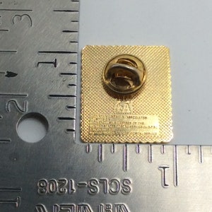 May include: A gold-colored square pin with a textured surface and a pin back. The pin has text that reads "L.A. & ASSOCIATES" and "OFFICIAL LICENSEE OF THE UNITED STATES POSTAL SERVICE (U.S.P.S.)".