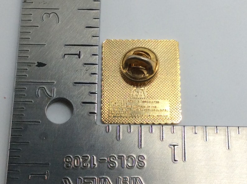 May include: A gold-colored square pin with a textured surface and a pin back. The pin has text that reads "L.A. & ASSOCIATES" and "OFFICIAL LICENSEE OF THE UNITED STATES POSTAL SERVICE (U.S.P.S.)".