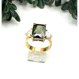 May include: A gold-toned ring featuring a large, rectangular green gemstone as the centerpiece, flanked by two smaller, pear-shaped clear stones. The ring is set on a clear display stand, showcasing its design and details.