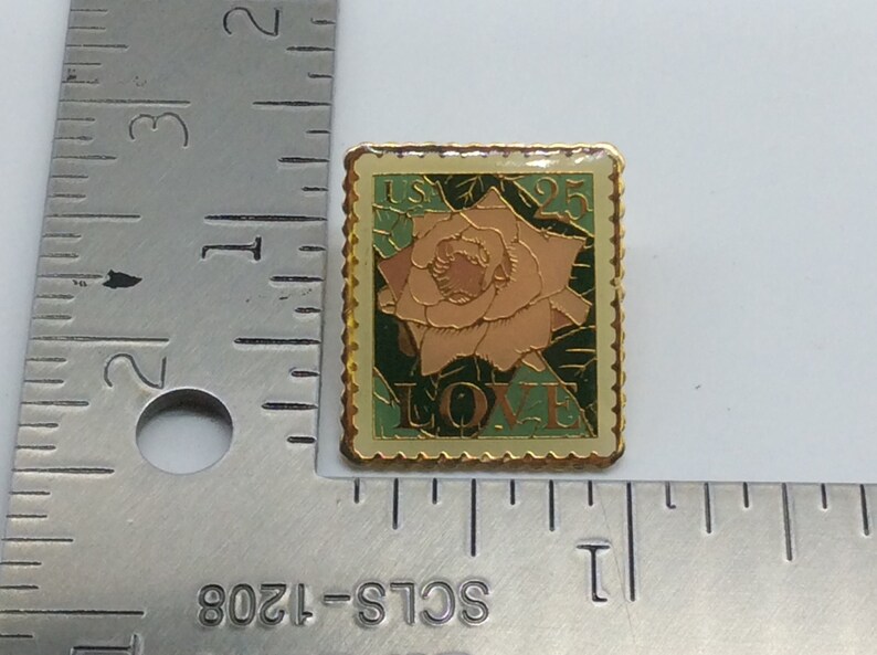May include: A rectangular pin with a gold border, featuring a rose design. The pin has a light green background with the text "USA 25 LOVE". The rose is peach-colored with green leaves.