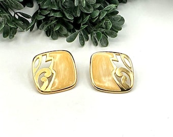 May include: A pair of square gold-toned earrings with a cream-colored enamel center and openwork design. The earrings have a swirling, geometric pattern and a polished finish. The background features green foliage.