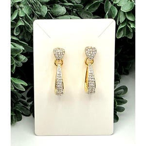 May include: A pair of gold-toned earrings with a heart-shaped top and a teardrop-shaped bottom, both encrusted with small, clear crystals. The earrings are displayed on a white card against a backdrop of green foliage.