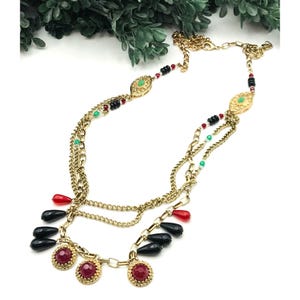 May include: A gold chain necklace with multiple strands and red, black, and green beads. The necklace has a pendant with a red stone and gold accents.