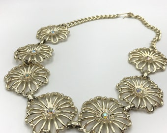 May include: Gold-toned necklace featuring openwork daisy-shaped pendants. Each daisy has a central iridescent crystal. The necklace is connected by a delicate chain and has a lobster clasp closure. A vintage-inspired jewelry piece.