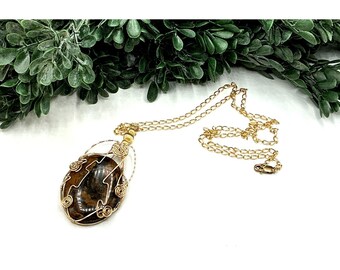May include: A gold-colored chain necklace with a large, oval pendant. The pendant features a brown and black stone wrapped in gold-colored wire. The necklace is displayed on a white surface with green foliage in the background.