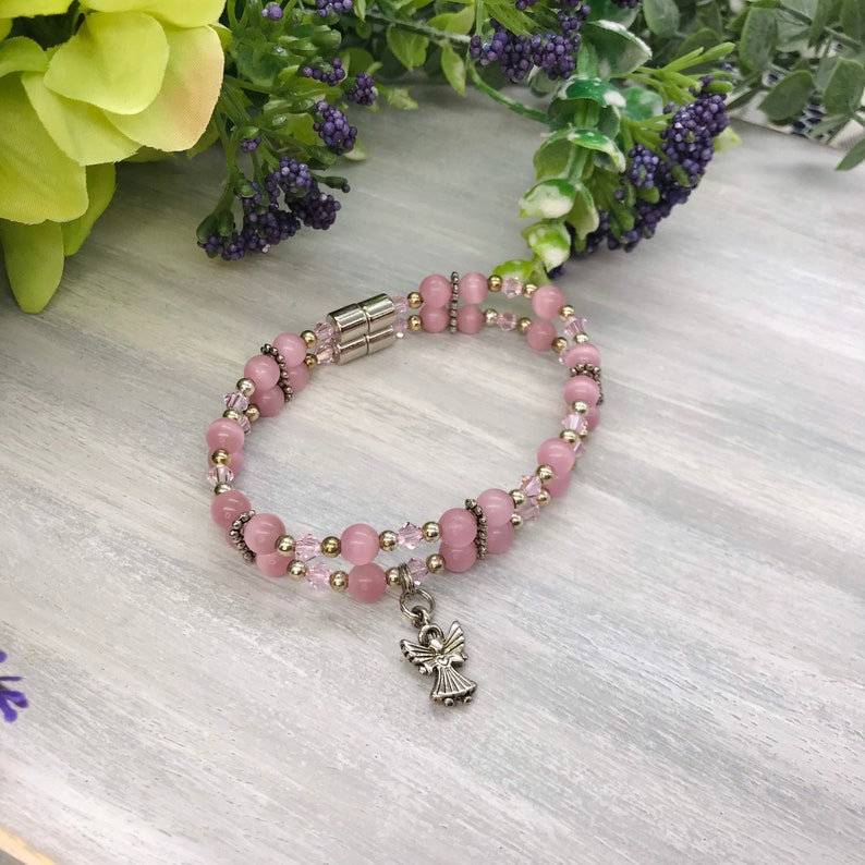 May include: A pink beaded bracelet with a silver angel charm. The bracelet is made with pink and gold beads and has a magnetic clasp.