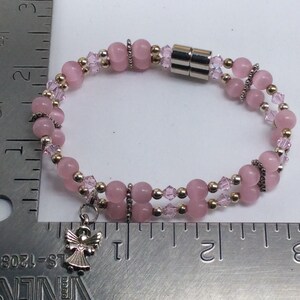 May include: A pink and gold beaded bracelet with a silver angel charm. The bracelet has a magnetic clasp and is approximately 7 inches long.