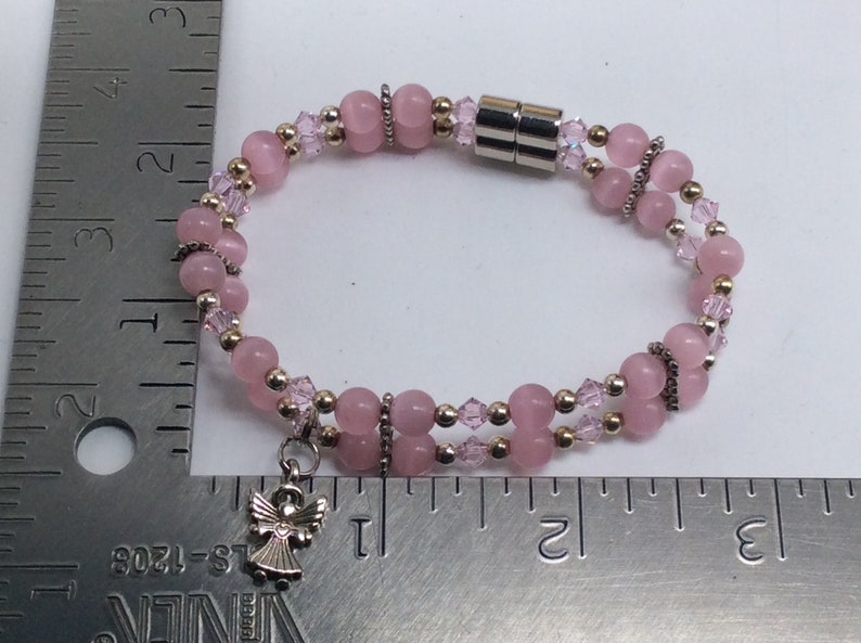 May include: A pink and gold beaded bracelet with a silver angel charm. The bracelet has a magnetic clasp and is approximately 7 inches long.
