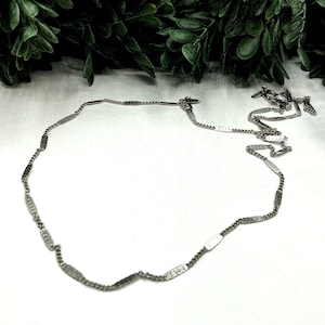 May include: A silver-toned necklace with a delicate chain and rectangular links. The necklace is displayed on a white surface with green foliage in the background. The chain is approximately 18 inches long.