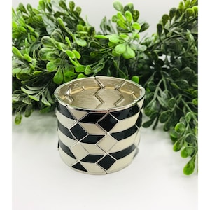 May include: A silver-toned stretch bracelet with a geometric design. The bracelet features black and off-white diamond and rectangular shapes. The bracelet is set against a backdrop of green foliage.