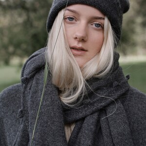 K&ouml;nnte beinhalten: Eine Frau tr&auml;gt eine graue Strickm&uuml;tze und einen grauen Strickschal. Der Schal ist um ihren Hals gewickelt und in einem lockeren Knoten gebunden. Ihr Haar ist lang und blond, und sie schaut in die Kamera.