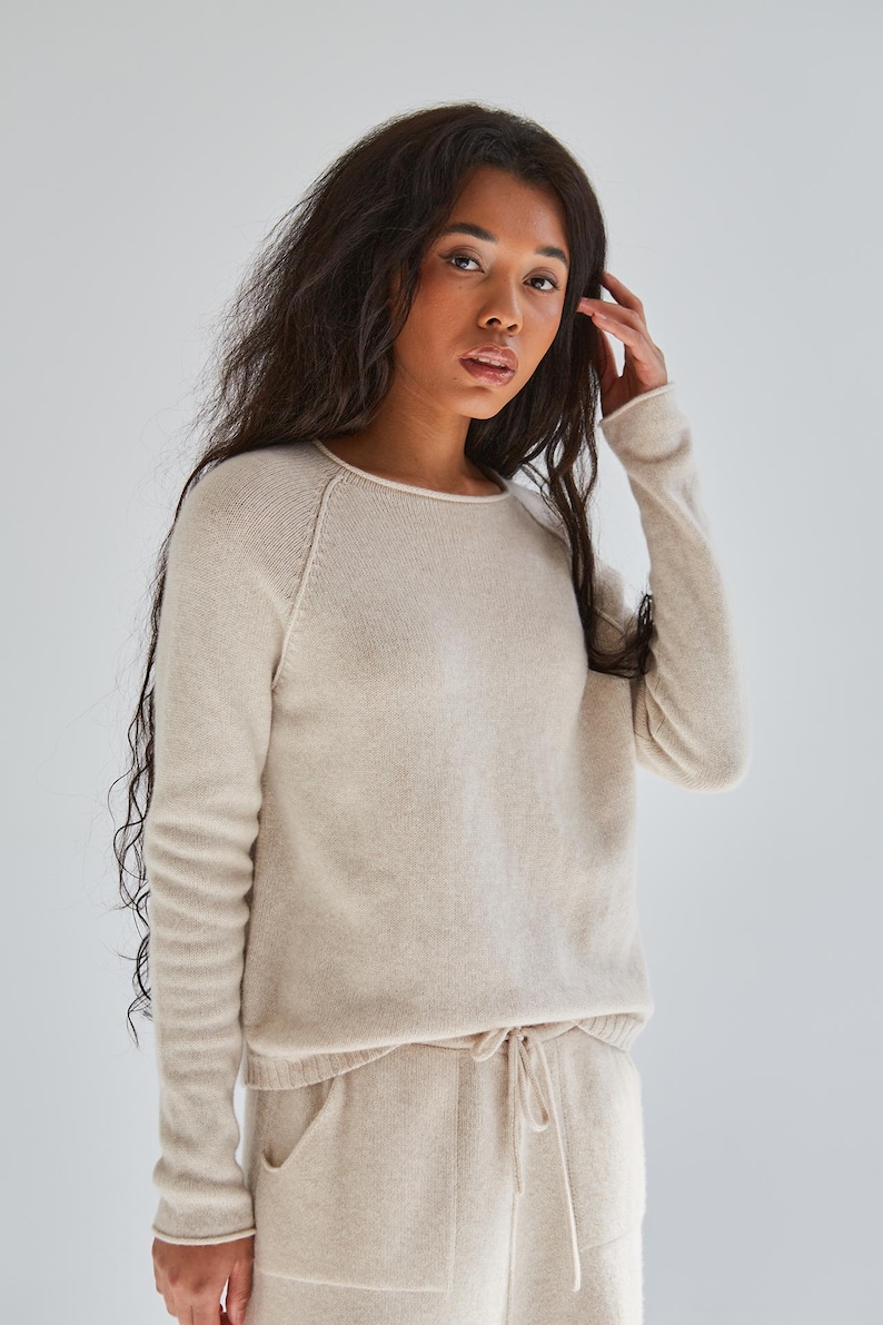 K&ouml;nnte beinhalten: Ein beigefarbener Langarmpullover mit Rundhalsausschnitt und Raglan&auml;rmeln, kombiniert mit einer passenden beigen Hose mit Kordelzug und zwei Vordertaschen. Das Set besteht aus einem weichen, fein gestrickten Stoff in einem hellen Ton.