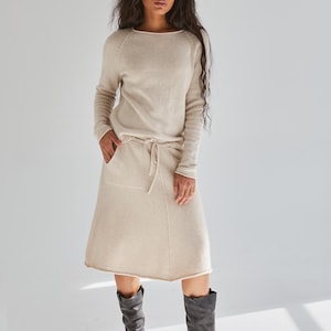 May include: A beige long-sleeved sweater with a round neckline and a matching knee-length skirt featuring a drawstring waist, two front pockets, and a raw hemline, paired with dark gray mid-calf leather boots with side buckle hardware.