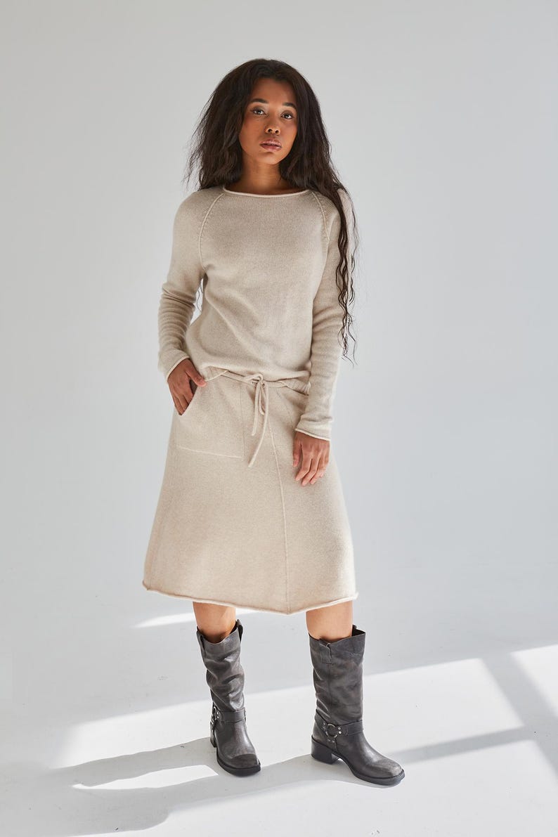 May include: A beige long-sleeved sweater with a round neckline and a matching knee-length skirt featuring a drawstring waist, two front pockets, and a raw hemline, paired with dark gray mid-calf leather boots with side buckle hardware.