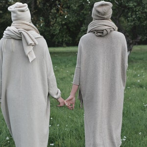 K&ouml;nnte beinhalten: Zwei Personen tragen lange, beige Strickpullover und passende Strickm&uuml;tzen und gehen nebeneinander auf einer grasbewachsenen Wiese.
