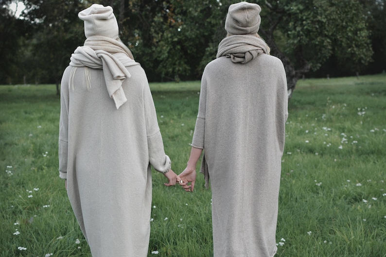 K&ouml;nnte beinhalten: Zwei Personen tragen lange, beige Strickpullover und passende Strickm&uuml;tzen und gehen nebeneinander auf einer grasbewachsenen Wiese.
