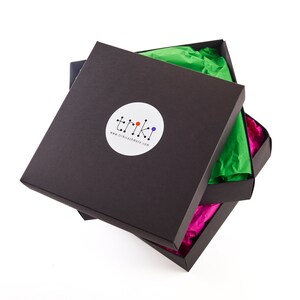 May include: Three black square boxes, partially open, revealing colorful tissue paper. The top box has a white circular sticker with the word "triki" and a colorful logo. The tissue paper is green and pink.