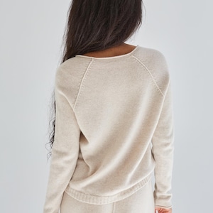 K&ouml;nnte beinhalten: Ein beigefarbener Strickpullover mit langen &Auml;rmeln, rundem Halsausschnitt und Raglan&auml;rmeln. Das Kleidungsst&uuml;ck ist aus einem weichen, fein gestrickten Material mit dezenter Textur gefertigt und besticht durch ein schlichtes, klares Design.