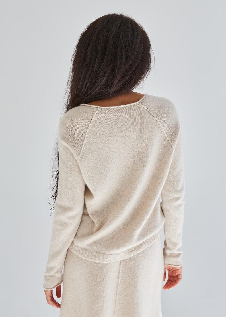 K&ouml;nnte beinhalten: Ein beigefarbener Strickpullover mit langen &Auml;rmeln, rundem Halsausschnitt und Raglan&auml;rmeln. Das Kleidungsst&uuml;ck ist aus einem weichen, fein gestrickten Material mit dezenter Textur gefertigt und besticht durch ein schlichtes, klares Design.
