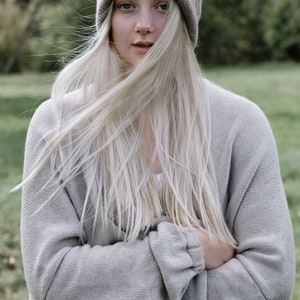K&ouml;nnte beinhalten: Eine Frau mit langen, blonden Haaren, die einen grauen Strickcardigan und eine graue Strickm&uuml;tze tr&auml;gt.