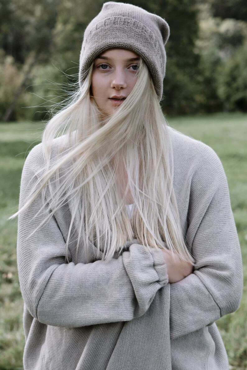 K&ouml;nnte beinhalten: Eine Frau mit langen, blonden Haaren, die einen grauen Strickcardigan und eine graue Strickm&uuml;tze tr&auml;gt.