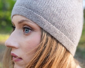 May include: A light beige knit beanie featuring a smooth stockinette stitch pattern on the crown and a textured garter stitch border along the bottom edge. The hat is crafted from a soft, fine-gauge yarn with a subtle heathered appearance.