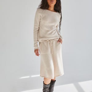 K&ouml;nnte beinhalten: Ein beigefarbener Pullover mit langen &Auml;rmeln und Rundhalsausschnitt sowie ein dazu passender knielanger Rock mit Kordelzug in der Taille und zwei Vordertaschen. Das Outfit wird mit dunkelgrauen wadenhohen Stiefeln mit seitlicher Schnalle kombiniert.