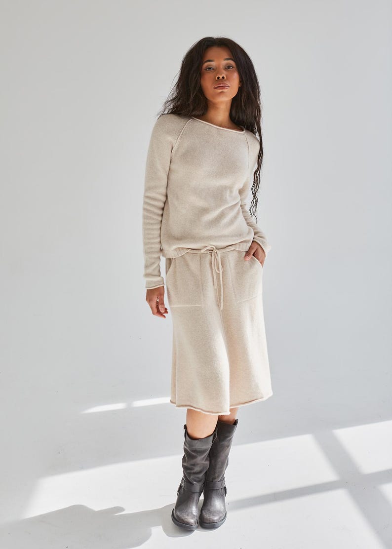 K&ouml;nnte beinhalten: Ein beigefarbener Pullover mit langen &Auml;rmeln und Rundhalsausschnitt sowie ein dazu passender knielanger Rock mit Kordelzug in der Taille und zwei Vordertaschen. Das Outfit wird mit dunkelgrauen wadenhohen Stiefeln mit seitlicher Schnalle kombiniert.