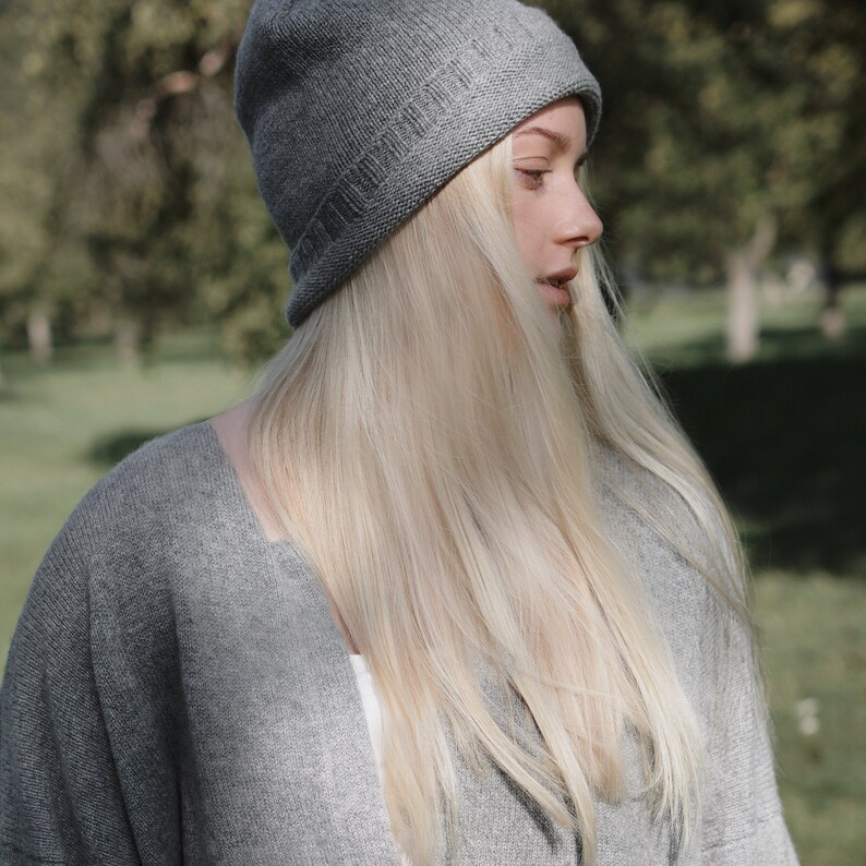 K&ouml;nnte beinhalten: Eine Frau mit langen blonden Haaren, die eine graue Strickm&uuml;tze und einen grauen Cardigan tr&auml;gt.