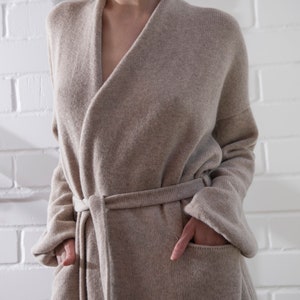 May include: A beige knit cardigan with a shawl collar, long sleeves, and a matching tie belt at the waist. The garment features two large front patch pockets and a textured knit construction, providing a relaxed fit that falls below the waistline.