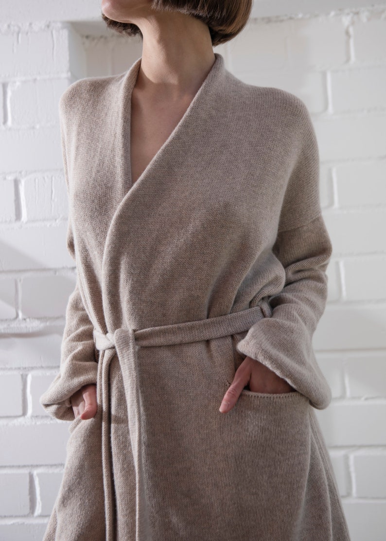 May include: A beige knit cardigan with a shawl collar, long sleeves, and a matching tie belt at the waist. The garment features two large front patch pockets and a textured knit construction, providing a relaxed fit that falls below the waistline.