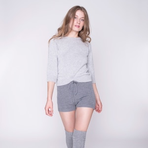 Könnte beinhalten: Ein hellgrauer Langarmpullover mit Rundhalsausschnitt kombiniert mit dunkelgrauen Strickshorts mit Kordelzug in der Taille und dazu passenden grauen Overknee-Socken. Das Set besteht aus einem weichen Strickstoff mit gleichmäßiger Textur.