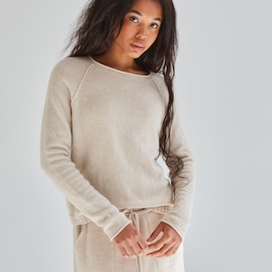 May include: A beige long-sleeved sweater with a round neckline and raglan sleeves, paired with a matching beige skirt featuring a drawstring waist and two front pockets. The set is crafted from a soft, fine-knit fabric with a consistent neutral tone.