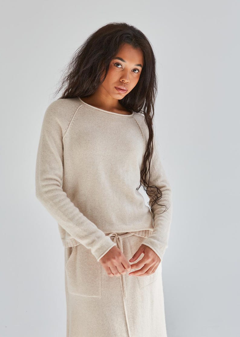 May include: A beige long-sleeved sweater with a round neckline and raglan sleeves, paired with a matching beige skirt featuring a drawstring waist and two front pockets. The set is crafted from a soft, fine-knit fabric with a consistent neutral tone.