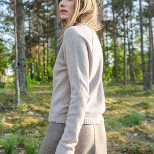 May include: A long-sleeved beige sweater with a round neckline and a ribbed hem, paired with a matching beige skirt. The sweater features a soft, knit texture and raglan sleeves, while the skirt has a smooth, structured appearance.