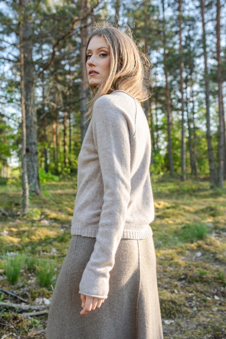 May include: A long-sleeved beige sweater with a round neckline and a ribbed hem, paired with a matching beige skirt. The sweater features a soft, knit texture and raglan sleeves, while the skirt has a smooth, structured appearance.