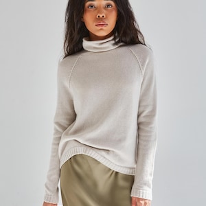 Könnte beinhalten: Ein langärmeliger, hellbeiger Rollkragenpullover mit geripptem Saum und Bündchen, kombiniert mit einem glatten, olivgrünen Midirock. Der Pullover hat Raglanärmel und eine weiche Strickstruktur, während der Rock einen dezenten, seidenartigen Glanz aufweist.