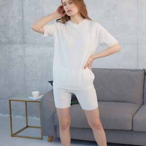 May include: A matching white knitwear set, consisting of a short-sleeved V-neck top and biker shorts. The outfit is made of a soft, ribbed material. The model is standing in a room with a gray sofa and a small side table.