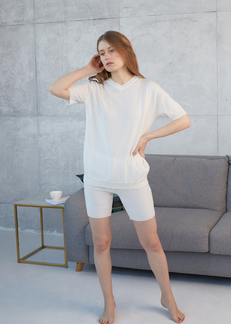 May include: A matching white knitwear set, consisting of a short-sleeved V-neck top and biker shorts. The outfit is made of a soft, ribbed material. The model is standing in a room with a gray sofa and a small side table.