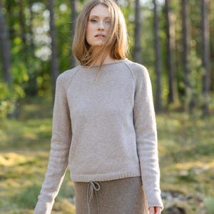 May include: A light beige long-sleeved sweater with a round neckline and raglan sleeves, paired with a darker brown skirt featuring a drawstring waist. The sweater has a fine knit texture, and the skirt is made of a solid, textured fabric.