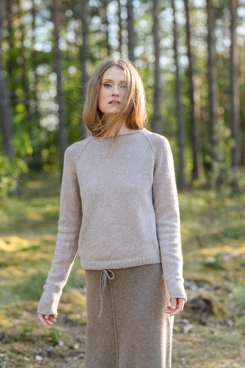 May include: A light beige long-sleeved sweater with a round neckline and raglan sleeves, paired with a darker brown skirt featuring a drawstring waist. The sweater has a fine knit texture, and the skirt is made of a solid, textured fabric.