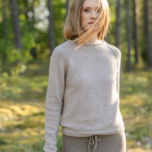 May include: A beige long-sleeved sweater with a round neckline and raglan sleeves, paired with a matching beige skirt featuring a drawstring waist. The knit garment has a soft, textured appearance with a ribbed hemline and simple construction.