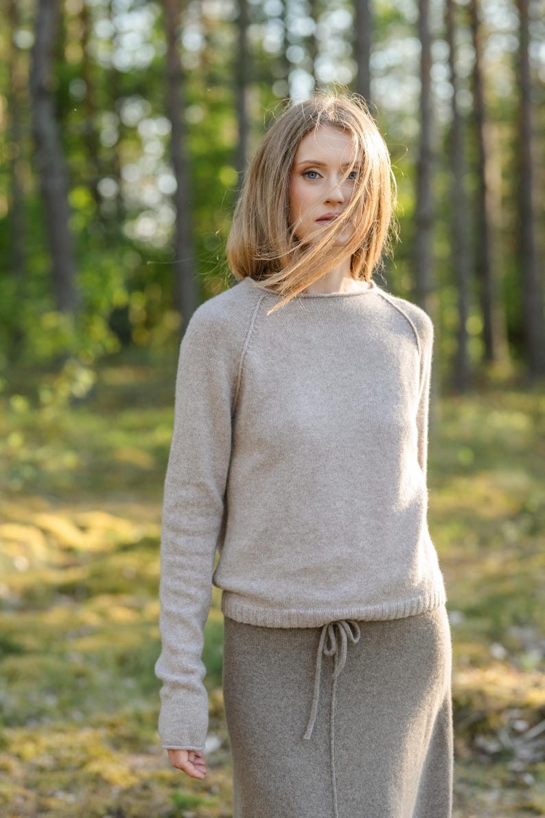 May include: A beige long-sleeved sweater with a round neckline and raglan sleeves, paired with a matching beige skirt featuring a drawstring waist. The knit garment has a soft, textured appearance with a ribbed hemline and simple construction.