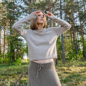 May include: A beige long-sleeved sweater with a crew neckline and a ribbed hem paired with a dark taupe ankle-length skirt featuring a drawstring waist and a vertical center seam. The sweater has raglan sleeves and a relaxed fit.