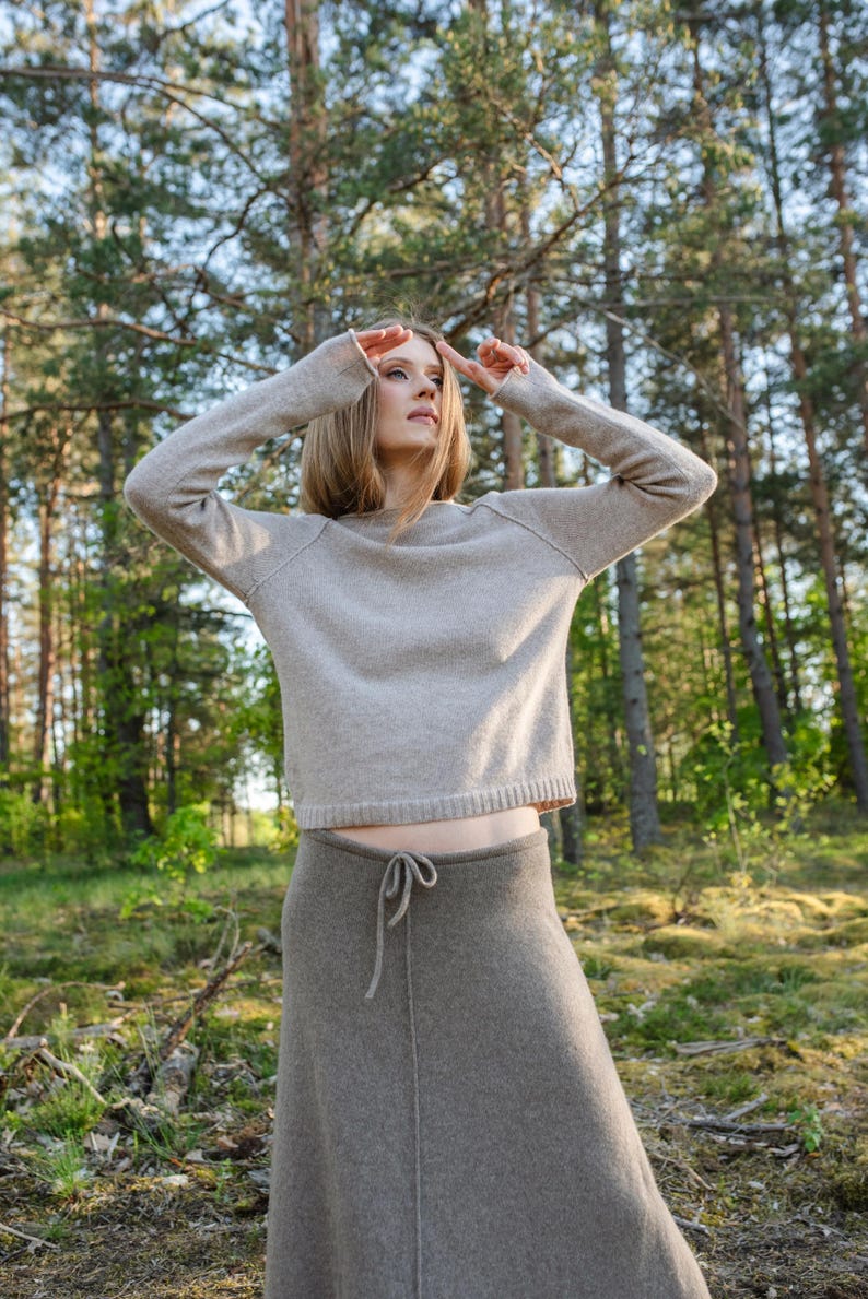 May include: A beige long-sleeved sweater with a crew neckline and a ribbed hem paired with a dark taupe ankle-length skirt featuring a drawstring waist and a vertical center seam. The sweater has raglan sleeves and a relaxed fit.