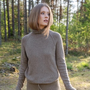 Könnte beinhalten: Ein langärmeliger taupefarbener Pullover mit hohem Rollkragen und Raglanärmeln, kombiniert mit einem passenden taupefarbenen Rock mit Kordelzug in der Taille. Das Set besteht aus einem weichen Strickstoff in einem einheitlichen, neutralen Farbton.