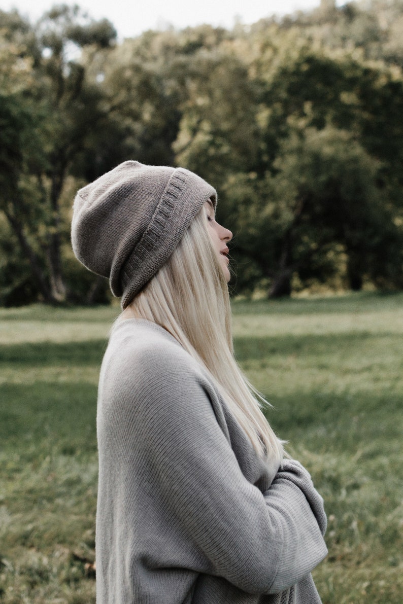 K&ouml;nnte beinhalten: Eine Frau mit langen blonden Haaren, die eine graue Strickm&uuml;tze und einen grauen Pullover tr&auml;gt. Sie steht auf einer gr&uuml;nen Wiese.