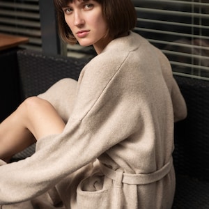 May include: A beige knit cardigan featuring long sleeves, a matching tie belt at the waist, and two front pockets. The garment is crafted from a soft, textured knit fabric with a relaxed fit, visible shoulder seams, and a clean, simple design.