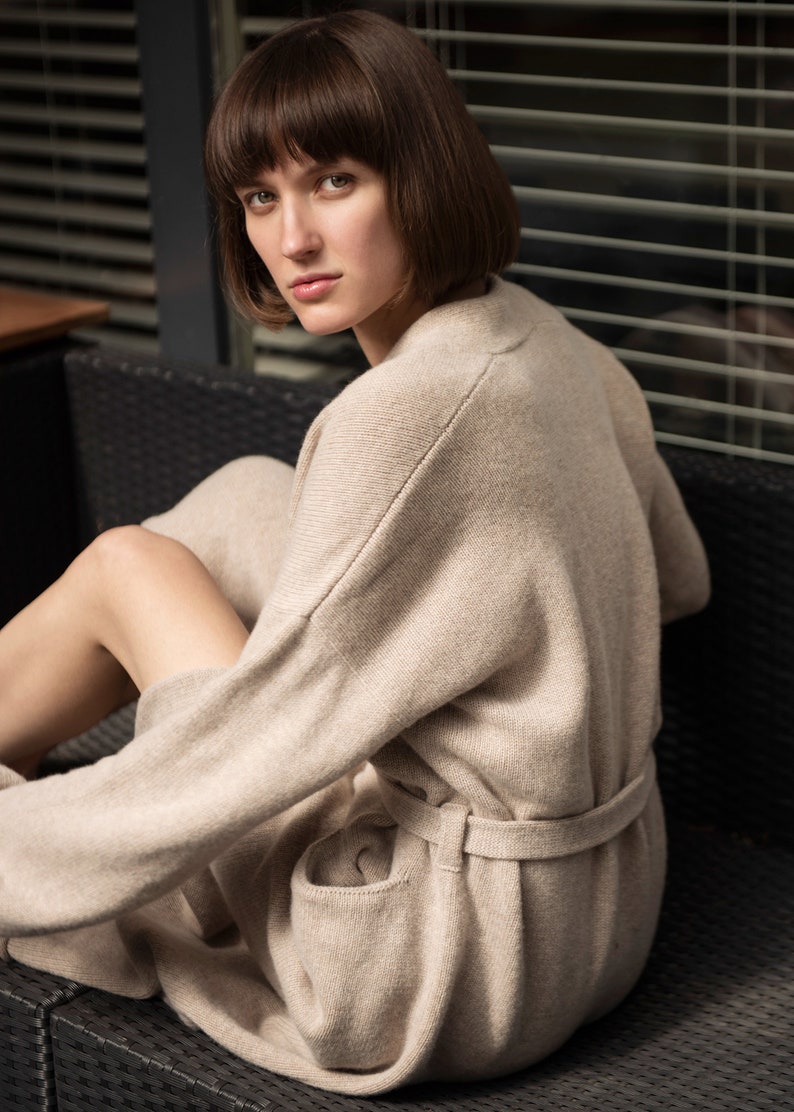May include: A beige knit cardigan featuring long sleeves, a matching tie belt at the waist, and two front pockets. The garment is crafted from a soft, textured knit fabric with a relaxed fit, visible shoulder seams, and a clean, simple design.