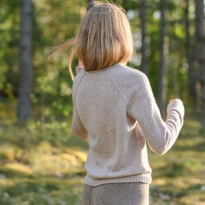 May include: A beige long-sleeved sweater with a round neckline and raglan sleeves, paired with a matching beige skirt. The sweater features a ribbed hem and cuffs, while the skirt has a visible vertical seam running down the center front.